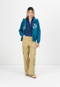 Young woman with blonde hair wearing blue GAP hoodie, dark blue shirt, beige cargo pants, and beige sneakers standing against white background.