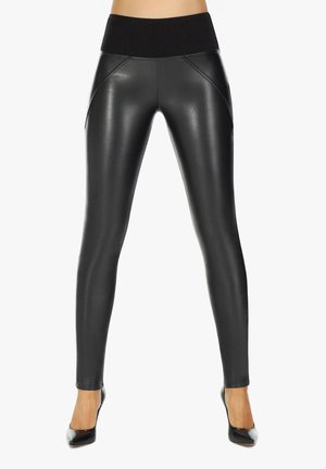 Kobieta ubrana w czarne leginsy ze sztucznej skóry z wysokim stanem i czarne szpilki z zaokrąglonymi noskami, stojąca z lekko rozstawionymi nogami.