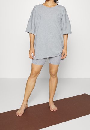 Person trägt lockeres graues T-Shirt und eng anliegende graue Shorts, steht barfuß auf brauner Yogamatte vor weißem Hintergrund.