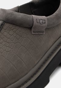 Chaussure gris foncé en daim texturé avec un profil élancé, un bout arrondi et une étiquette logo UGG. Dispose d'une semelle noire contrastante.
