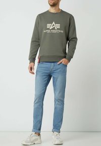 Felpa verde oliva con logo bianco, scollo a giro, polsini a costine, abbinata a jeans slim-fit azzurri e sneakers bianche.