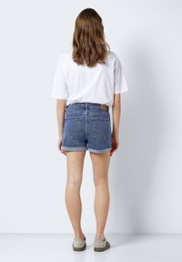 Pantalones cortos de mezclilla en azul, con una cintura alta, dobladillos en el extremo y bolsillos traseros. Combinados con una camiseta blanca holgada y zapatos grises.