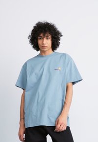 Light blue cotton t-shirt with short sleeves, crew neckline, and small embroidered sun logo on the chest. Smooth fabric texture.