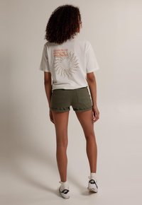 Camiseta blanca de algodón con un gráfico de sol y texto en la parte posterior, combinada con pantalones cortos de mezclilla verde oliva y zapatillas blancas con acentos negros.