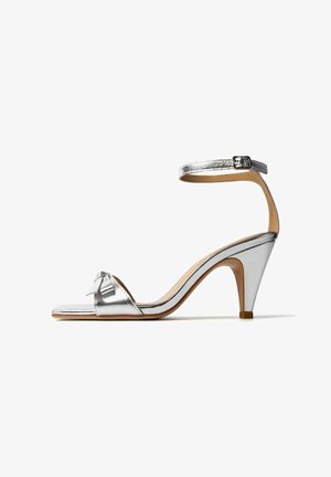 Sandalo argento con tacco alto, cinturino alla caviglia, punta aperta e fiocco decorativo sulla fascia frontale, mostrato su sfondo bianco.