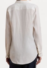Persona que lleva una camisa de botones de manga larga color beige claro con puños, vista desde atrás, combinada con pantalones oscuros.