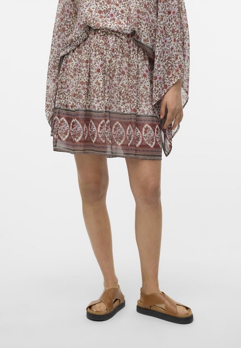 Jambes portant des sandales en cuir marron et une jupe ample à motif floral avec un bord rouge et blanc.