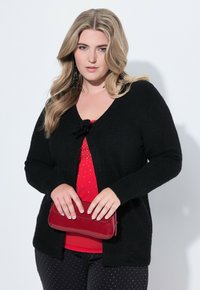 Žena s dlouhými blond vlasy má na sobě černý cardigan přes červený top, drží malou červenou kabelku a má na sobě černé kalhoty s bílými tečkami.