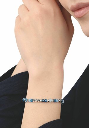 Nadgarstek z bransoletką z koralików w naprzemiennych kolorach: niebieskim, srebrnym i jasnoniebieskim, na nadgarstku osoby na białym tle.