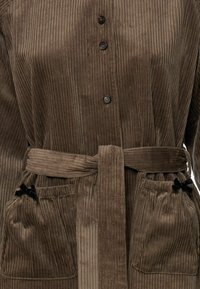 Barna kordszövetből készült gombos felső, amelyhez kötött öv és két elülső zseb tartozik, ezeket kis fekete masnik díszítik.