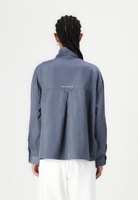 Blå lett button-up skjorte med bakfeller, brodert "BLANCHE"-logo, og justerbare mansjetter. Vist på en blank bakgrunn.
