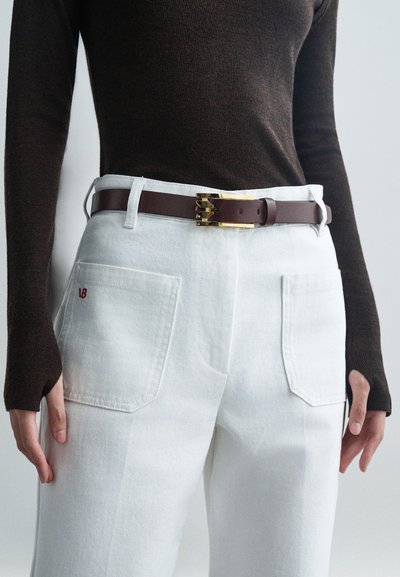 Pantaloni din denim alb cu două buzunare pe spate și un logo roșu. Asortați cu o curea din piele maro cu o cataramă auriu și un top ajustat de culoare maro închis.