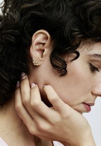 Orecchino a perno a forma di nodo dorato con gemme trasparenti incorporate, di forma circolare, superficie testurizzata, indossato da un modello con capelli scuri e arruffati.