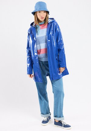 Femme portant un chapeau seau bleu, un imperméable bleu brillant, un pull rayé, un jean retroussé, des baskets bleues et un foulard autour du cou, debout sur un fond blanc.