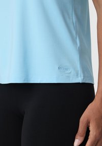 Světle modrý top s krátkým rukávem vyrobený z měkké tkaniny, s přímým lemem a nenápadným logem na spodní levé straně. Spárováno s černými legínami.