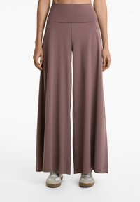 Vida, högmidjade byxor i mauve, tillverkade av mjukt tyg. Har en slät midjeband och en flödande silhuett. Visas med ljusa sneakers.