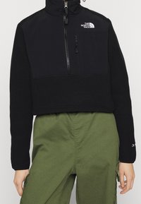 Svart kort puffa jacka med dragkedja fram, fleece-textur, hög krage och logotypdetalj på bröstet. Kombinerad med gröna cargobyxor.