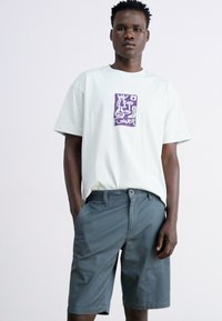 Tricou verde deschis cu design grafic purpuriu; asociat cu pantaloni scurți gri. Tricoul are o croială lejeră; pantalonii scurți au închidere cu nasture și buzunare.