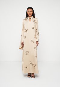 Beige hosszú ruha virágos hímzéssel, kulcslyuk kivágással és kontrasztos gallérral. Hosszú, laza ujjaival és sima textúrájával rendelkezik.