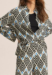 Conjunto de ropa negro y beige con un patrón que presenta diseños geométricos con detalles en azul. Camiseta holgada con cuello y cierre de botones, pantalones a juego.