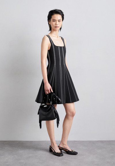 Robe sans manches noire avec des clous blancs verticaux, jupe évasée et décolleté carré. Associée à un sac à main noir et des talons ouverts.