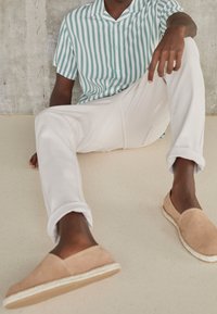 Gestreept T-shirt met korte mouwen in groen en wit, gecombineerd met witte broek en beige espadrilles, zittend op een glad, lichtgekleurde ondergrond.