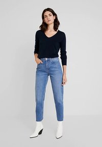 Marineblauwe trui met V-hals, lichtblauwe jeans met hoge taille en witte enkellaarsjes met hak. Gladde texturen; nauwsluitend model met taps toelopende pijpen.