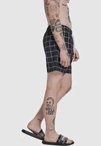 Zwarte zwembroek met een wit ruitpatroon, taille met trekkoord en contrasterende zijnaden. Getatoeëerd been zichtbaar, gecombineerd met zwarte sandalen.