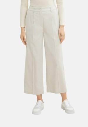 Pantalons larges, gris clair avec un devant plissé, fabriqués dans un tissu texturé. Associés à des baskets blanches pour un look décontracté.