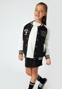 Giacca varsity bianca e nera con dettagli ricamati e bottoni a pressione, abbinata a una camicia bianca, una gonna nera e calze al ginocchio.