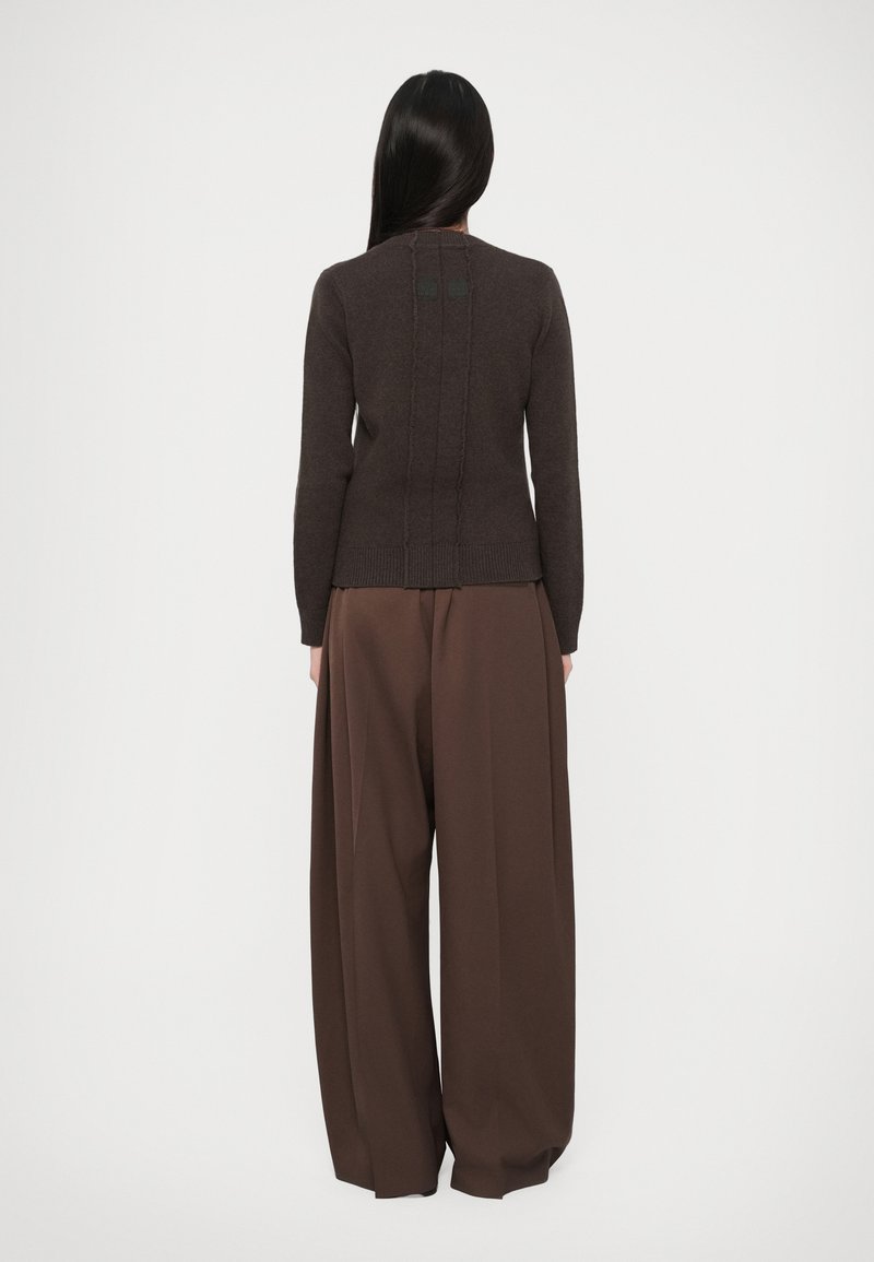 Woman with long dark hair wearing a dark brown fitted sweater and wide-legged brown trousers standing against a plain background.