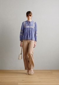Blåmønstret bluse med lange ærmer, beige vide bukser, kombinert med klare solbriller og en hvit håndveske, på et tregulv.