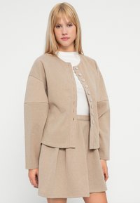 Jachetă textured bej cu guler rotund, închidere frontală cu nasturi și mâneci largi, asortată cu o fustă corespunzătoare.