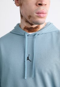 Sudadera azul claro de tela suave con cordones, con un logo negro de Jordan en el pecho. Acercamiento al cuello y detalles faciales.