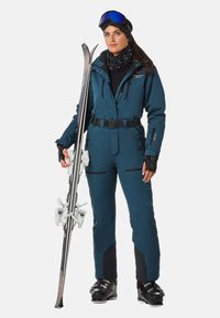 Skianzug in Türkis, aus wasserabweisendem Stoff gefertigt. Verfügt über einen Reißverschluss vorne, einen verstellbaren Gürtel und verstärkte Akzente. Hält Skier.