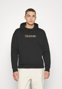 Felpa nera in misto cotone con tasca a marsupio frontale, cappuccio con cordino e logo dorato "Calvin Klein" sul petto.