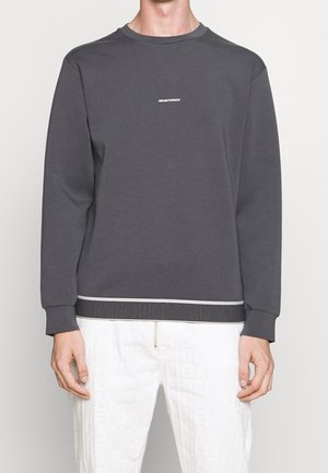 Man draagt een donkergrijze trui met lange mouwen met kleine witte tekst en witte broek met subtiel patroon, staand tegen een effen achtergrond.