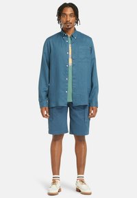 Camisa azul de botões com mangas longas, um bolso no peito, e t-shirt gráfica verde por baixo. Combinada com calções azuis a condizer e sapatos brancos.