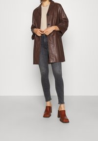 Osoba ubrana w brązowy skórzany płaszcz, beżowy sweter, ciemnoszare dopasowane dżinsy oraz brązowe czółenka na obcasie, stojąca na jednolitym tle.