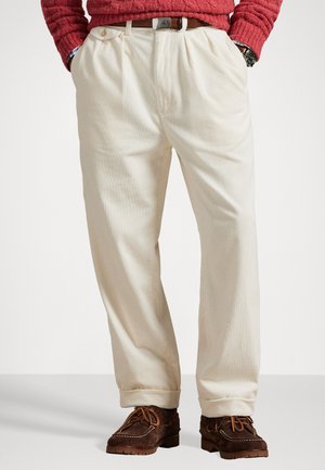 Pantalones de pana beige con un corte relajado, que presentan un frente plisado, bolsillos laterales y un dobladillo con vuelta, combinados con zapatos de ante marrones.