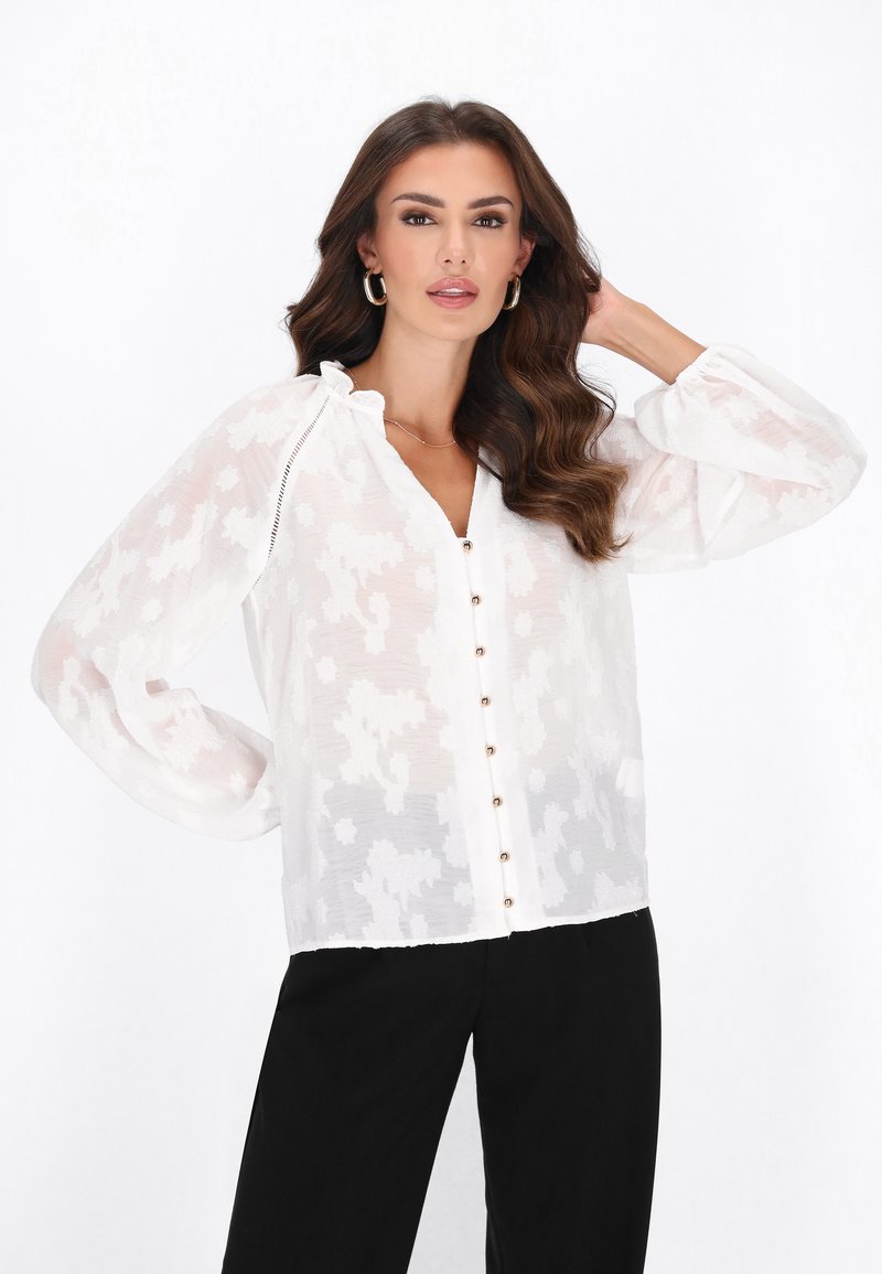 Blusa blanca transparente con patrón texturizado, con cierre de botones al frente, cuello con volantes y mangas largas. Combinada con pantalones negros.
