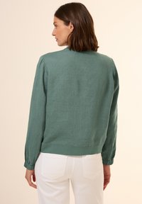 Groene linnen blazer met een ronde nek, elastische manchetten en een iets verlaagd zoom. Gecombineerd met witte broek voor contrast.