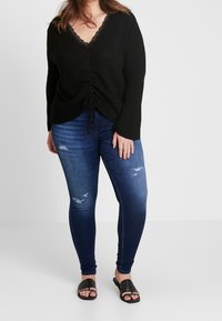 Suéter negro de canalé con encaje en el borde, detalle fruncido en la cintura, combinado con jeans ajustados azul oscuro con toques desgastados y sandalias planas.
