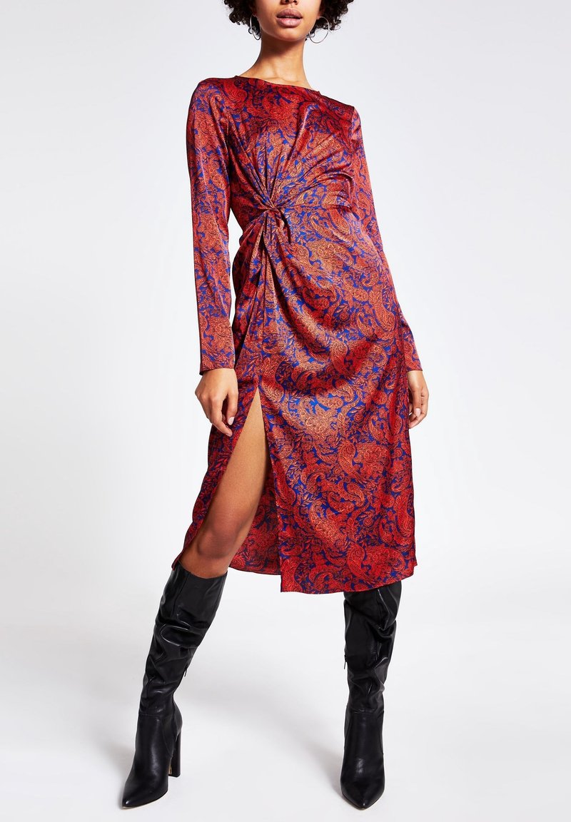 Femme portant une robe à manches longues à motifs floraux rouge et bleu avec une fente latérale et des bottes noires mi-mollet à talons.