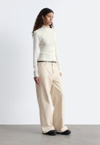 Langärmliges, elfenbeinfarbenes Oberteil mit geripptem Textur und Rüschenkragen, kombiniert mit weiten beigen Cordhosen und klobigen schwarzen Schuhen.