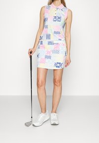 Krageklänning utan ärmar med patchworkdesign i pastellfärger, som visar olika mönster. I kombination med vita sportskor och en golfklubb.