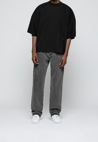 Persona vistiendo una camiseta negra extragrande, jeans gris oscuro holgados y zapatillas blancas, de pie contra un fondo claro y liso.