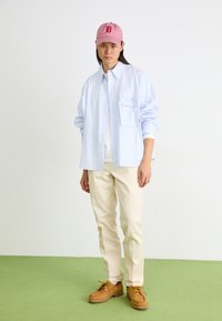 Cămașă button-up cu dungi albastre deschise, tricou alb dedesubt, pantaloni crem, pantofi maro cu șireți galbeni, șapcă roz cu logo.