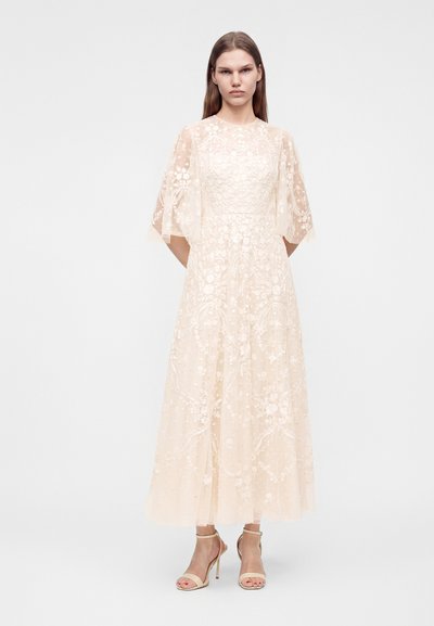 Femme debout portant une longue robe crème transparente avec broderies florales et manches courtes évasées, associée à des sandales à talons beiges.