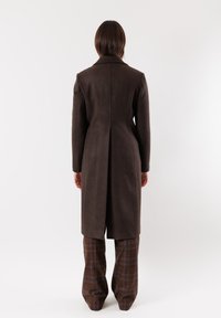 Cappotto di lana marrone con colletto e spacco posteriore, abbinato a pantaloni a gamba larga a quadri nelle tonalità di marrone e blu, visto da dietro.